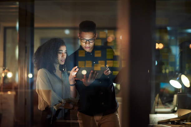 Two coworkers stand at a glass wall, looking at a tablet with sticky notes behind them in a dimly lit office.”,
