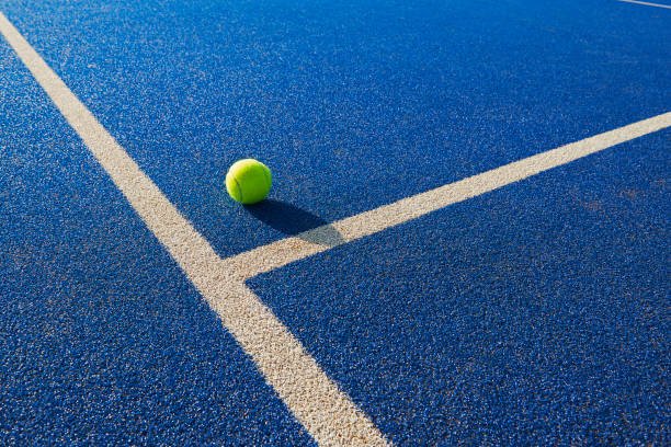 Tennis ball resting on a blue court at the intersection of white boundary lines, casting a shadow on the surface.