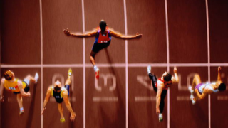 Overhead view of eight sprinters leaping midair from the track, arms outstretched as they race in colored uniforms on a red lane-lined track.
