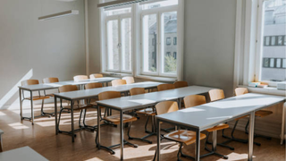 Empty classroom with rows of wooden desks and chairs, bright natural light from large windows on white walls.